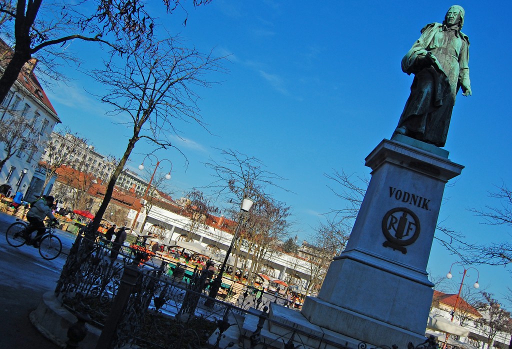 Vodnik is turning his back on both the Ljubljana market and the Dragon Bridge, instead facing the castle.